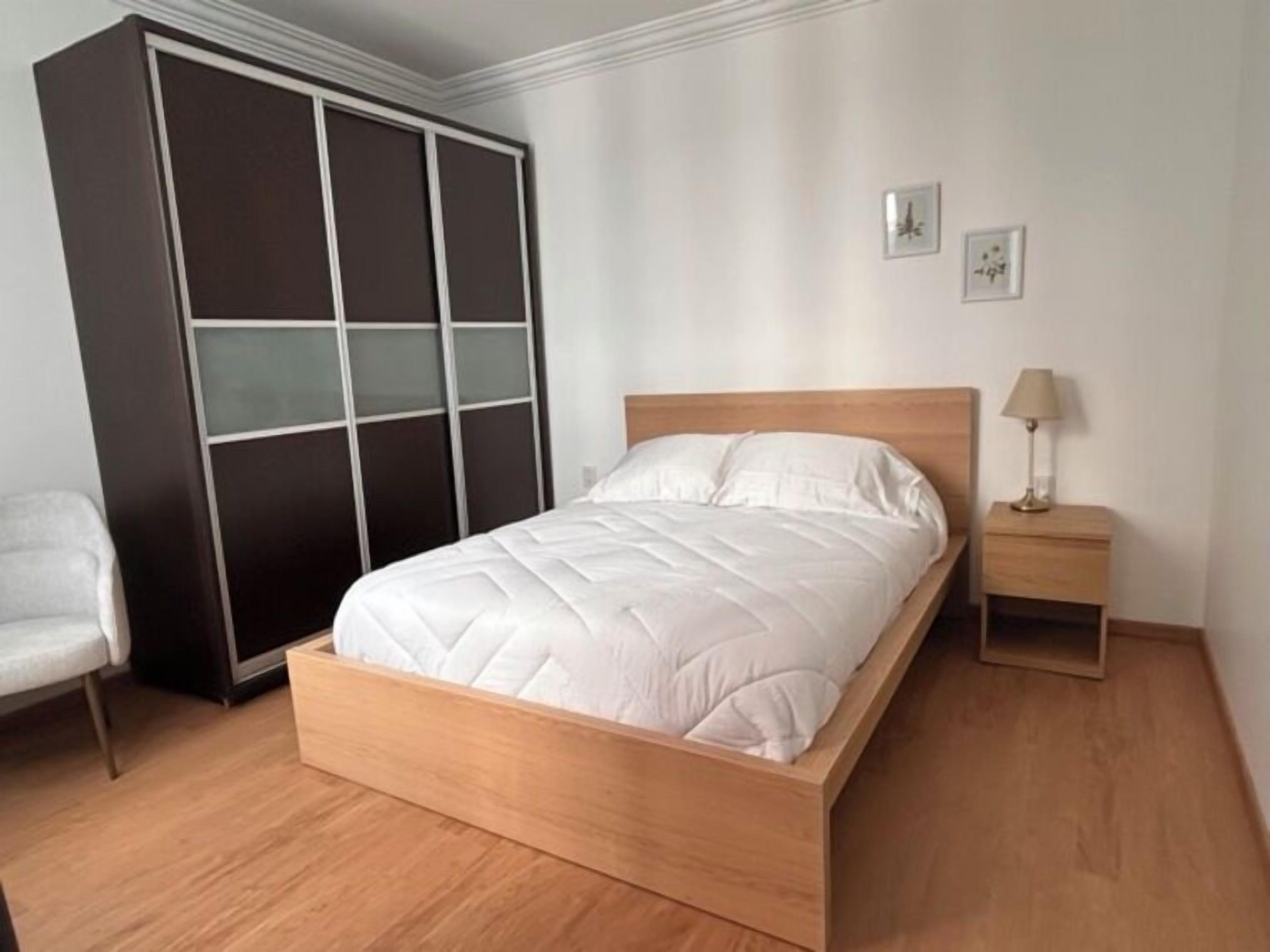 A bedroom with a wooden bed, a nightstand, a chair, and a wardrobe with frosted glass panels.