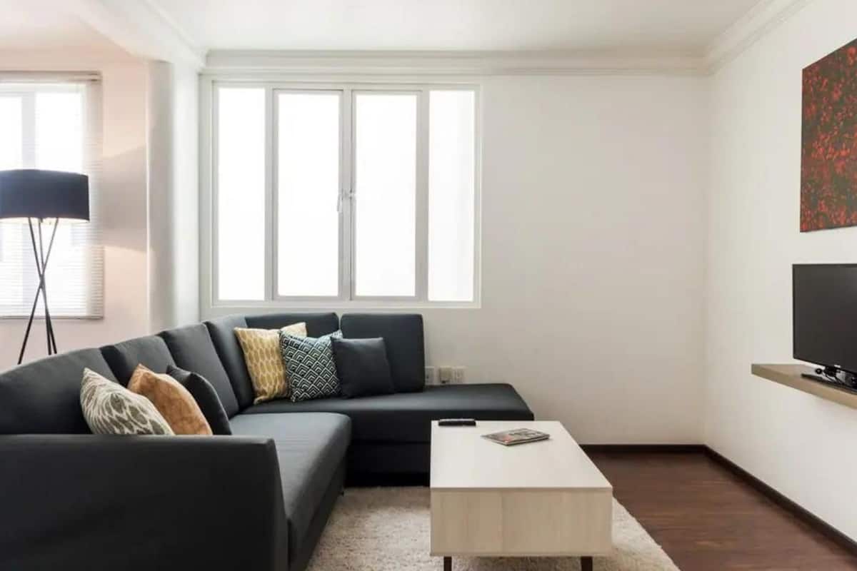 Ein modernes Wohnzimmer mit einem Ecksofa, einem Couchtisch, einem Flachbildfernseher auf einem Holzboden und großen Fenstern mit Jalousien.