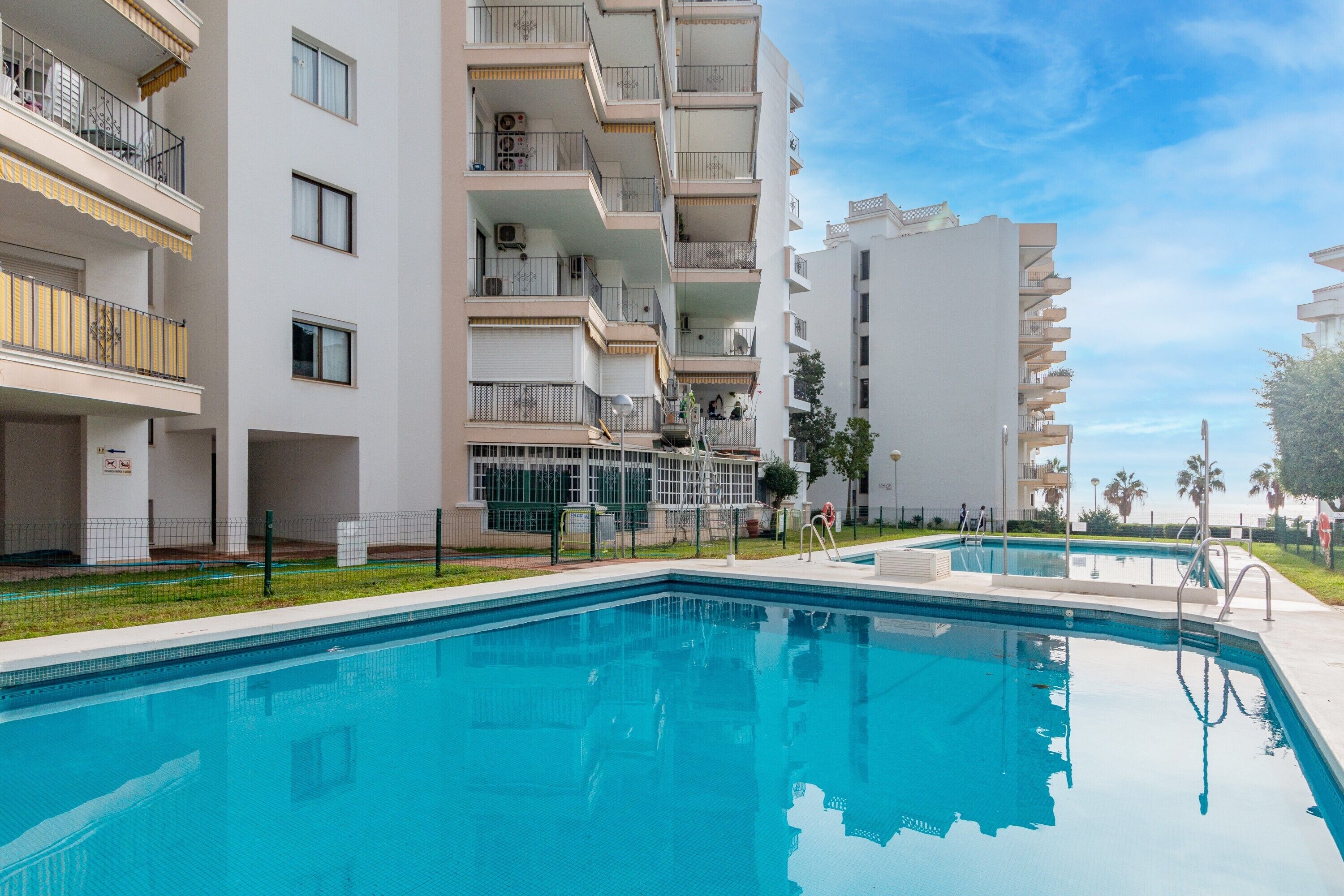 A swimming pool with a clear blue water surface, surrounded by white multi-story buildings with balconies and a clear sky above.