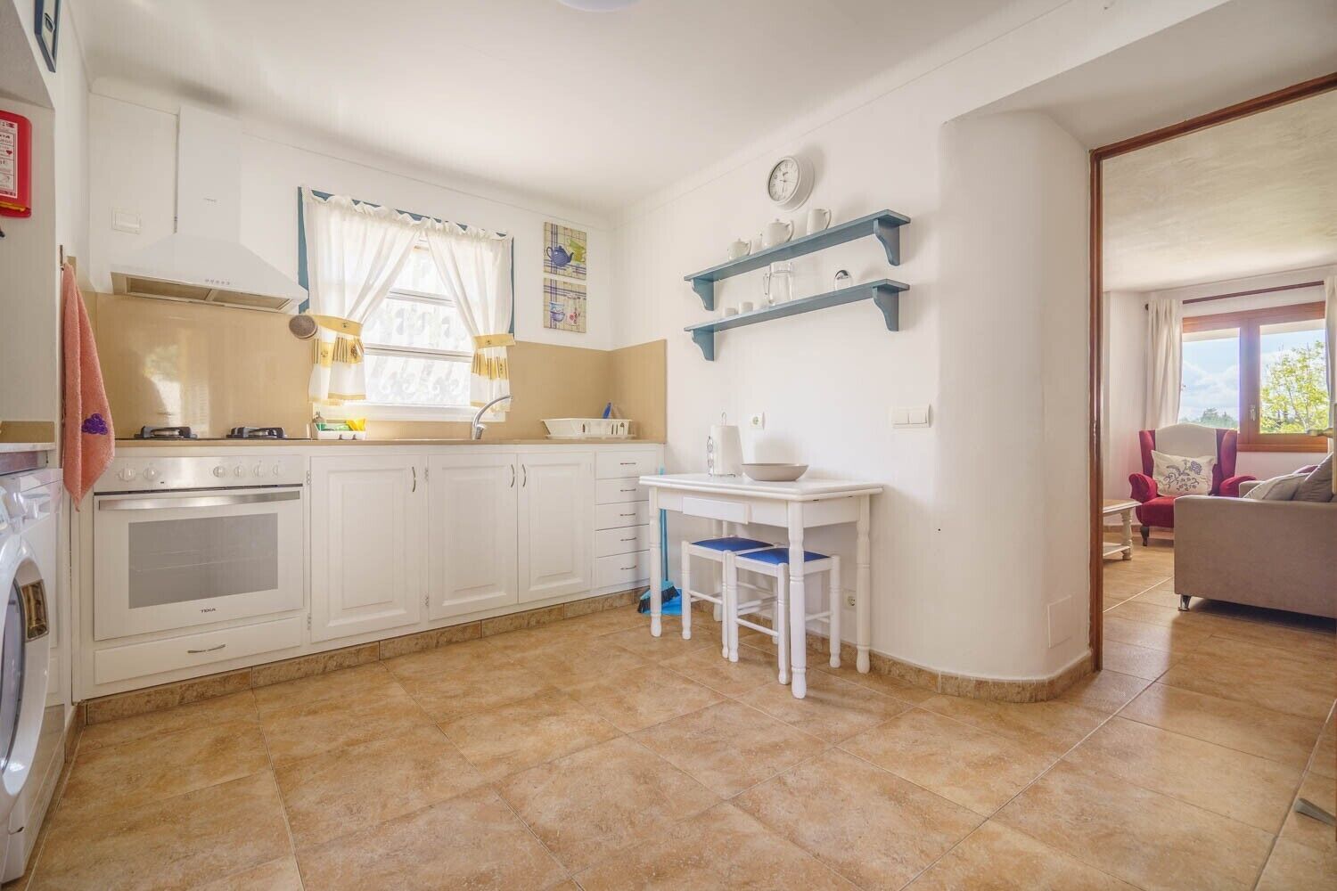 Una cocina con armarios blancos, una estufa y una mesa de comedor con sillas. Hay una ventana con cortinas y un reloj en la pared.