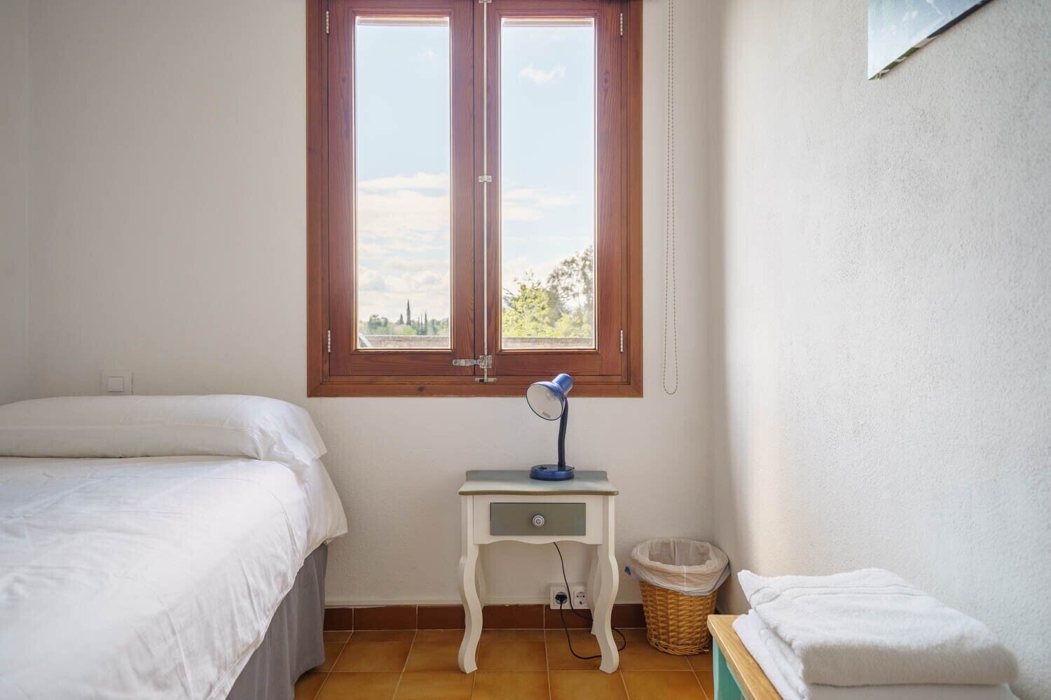 A bedroom with a window, a bedside table with a lamp, a small trash bin, and a bed.