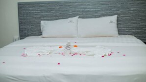 A neatly made bed with white linens and pillows, a towel arranged on the bed, and scattered rose petals.