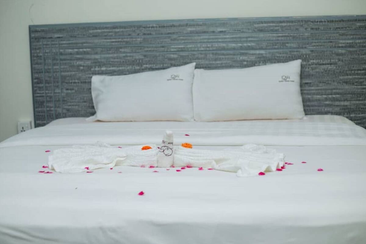 A neatly made bed with white linens and pillows, a towel arranged on the bed, and scattered rose petals.