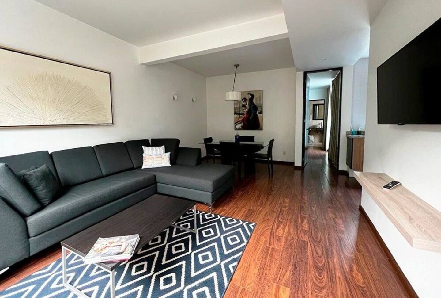 A modern living room with a sectional sofa, a dining area with a table and chairs, a patterned rug, and a flat-screen TV mounted on the wall.