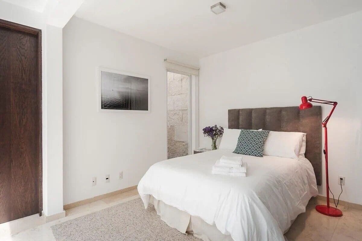 Un dormitorio con una cama, una lámpara roja de pie, un cuadro en la pared y una puerta.
