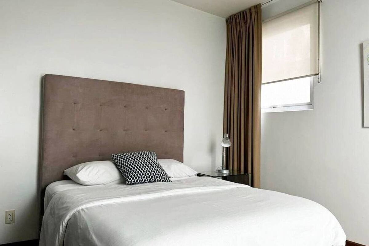 Una cama bien hecha con cabecera, una lámpara de noche y una ventana con cortinas.