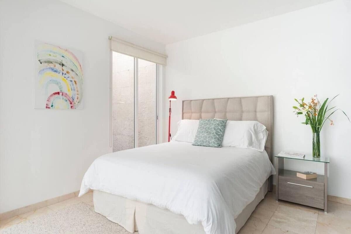 Un dormitorio con cama, mesita de noche, lámpara y una obra de arte en la pared.