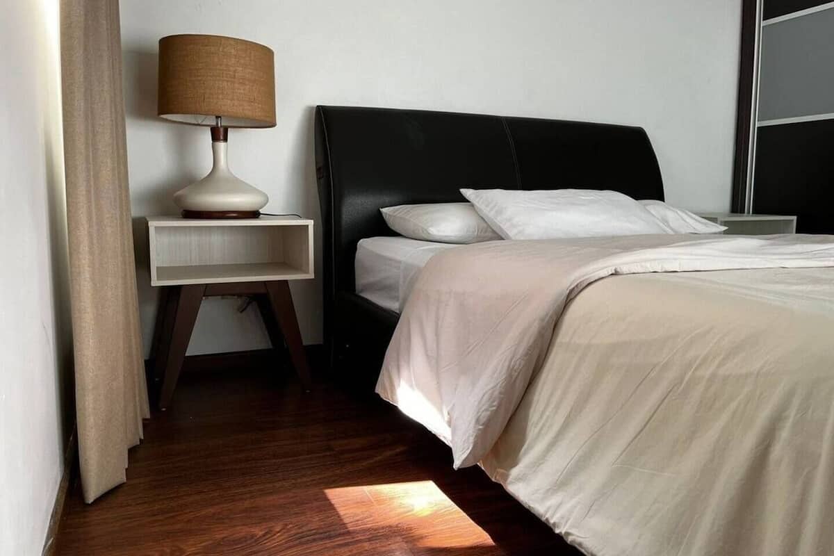 Una cama bien hecha con sábanas blancas, cabecera negra y una lámpara de noche sobre una mesita de madera.