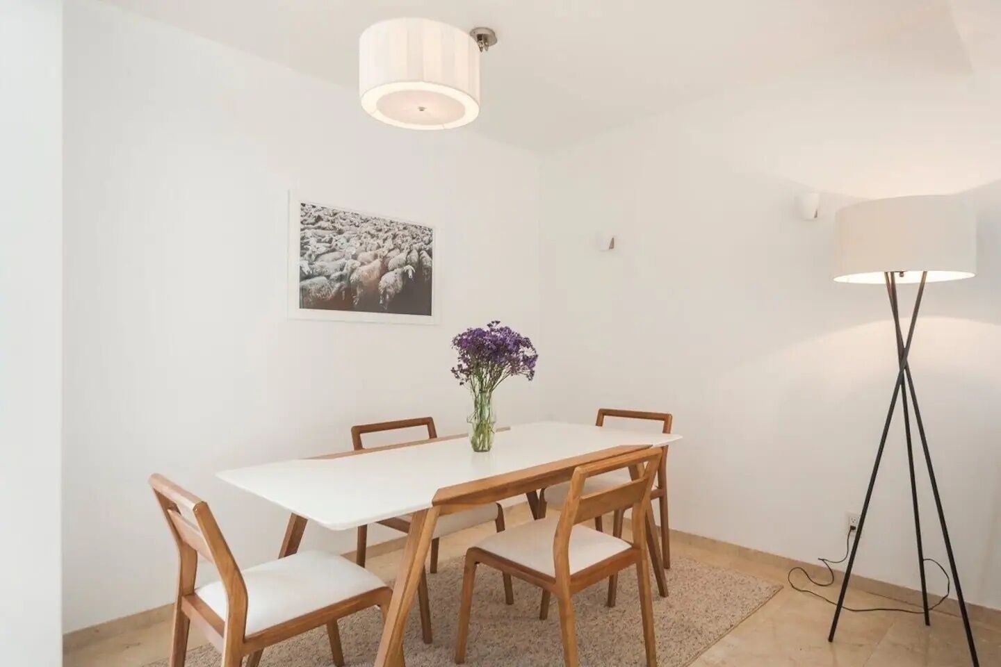 Ein Essbereich mit einem weißen Tisch, Holzstühlen, einer Vase mit violetten Blumen, einem Wandbild und einer Stehlampe.
