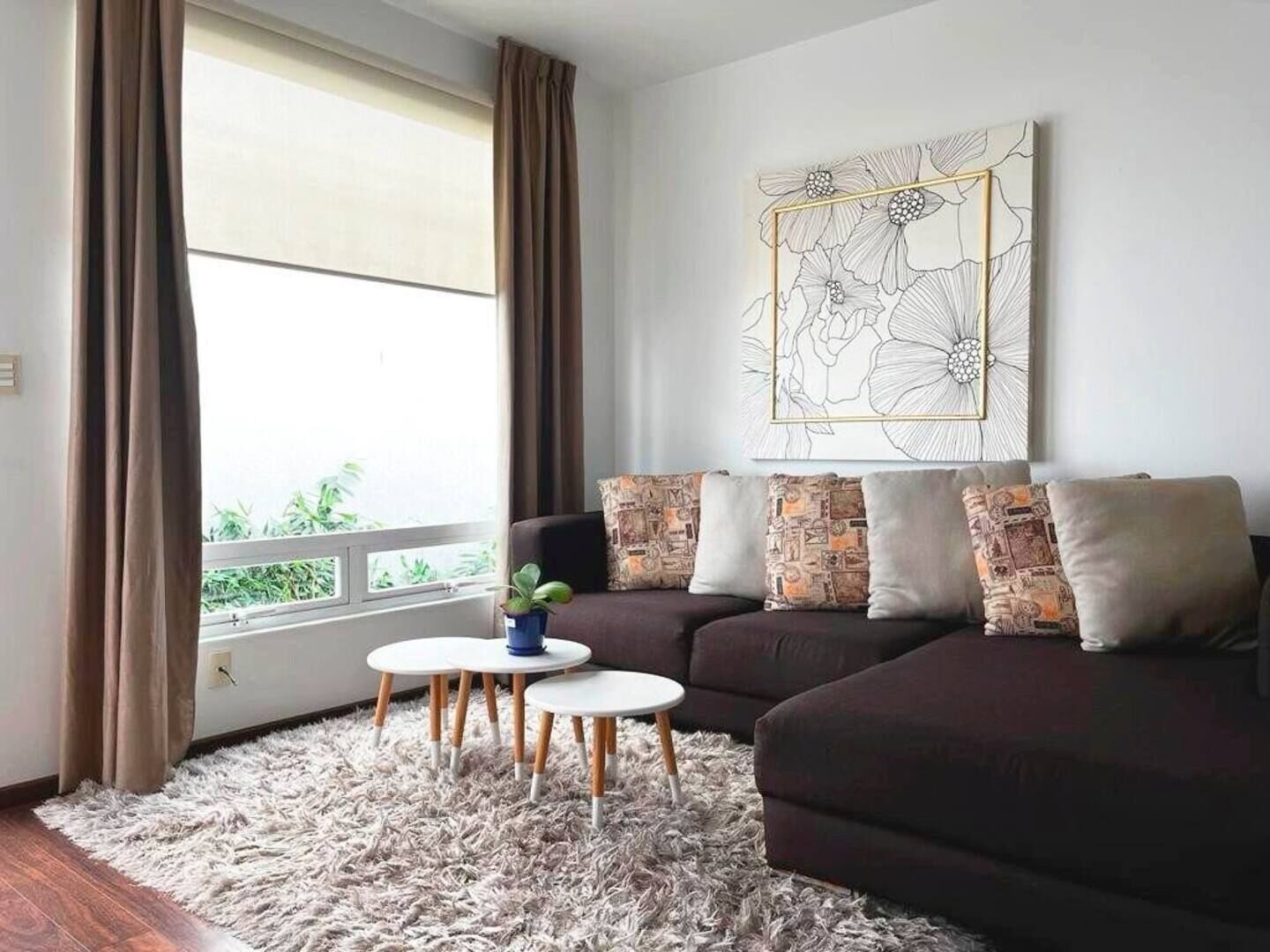 A living room with a dark sofa, patterned cushions, a fluffy rug, and a large window with curtains.