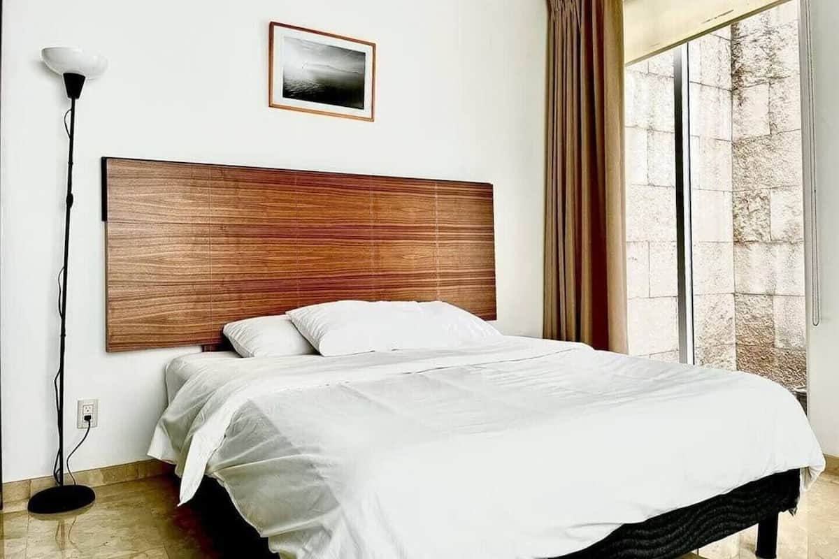 Un dormitorio con cabecera de madera, cama con ropa de cama blanca, una lámpara de pie y una ventana con cortinas.