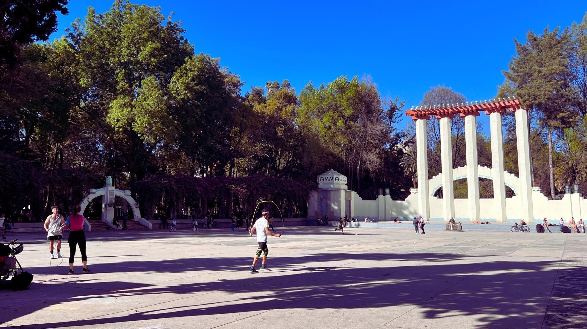 An open plaza with a large white structure featuring red pillars, surrounded by trees and people engaging in various activities.