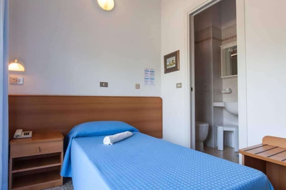 A small, neatly arranged bedroom with a single bed, a bedside table, a nightstand, and a bathroom visible through an open door.