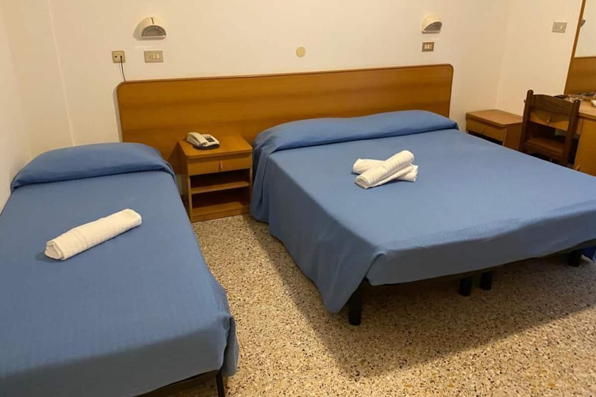 A hotel room with two single beds, each with a blue bedspread and a rolled white towel. There is a wooden headboard, a bedside table with a lamp, and a small wooden chair.