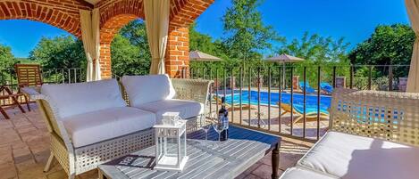 An outdoor seating area with a pool view, featuring a white sofa, wicker chairs, and a table with a bottle and glass.