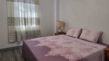 A bedroom with a bed, a ceiling fan, a nightstand, a lamp, and a window with curtains.