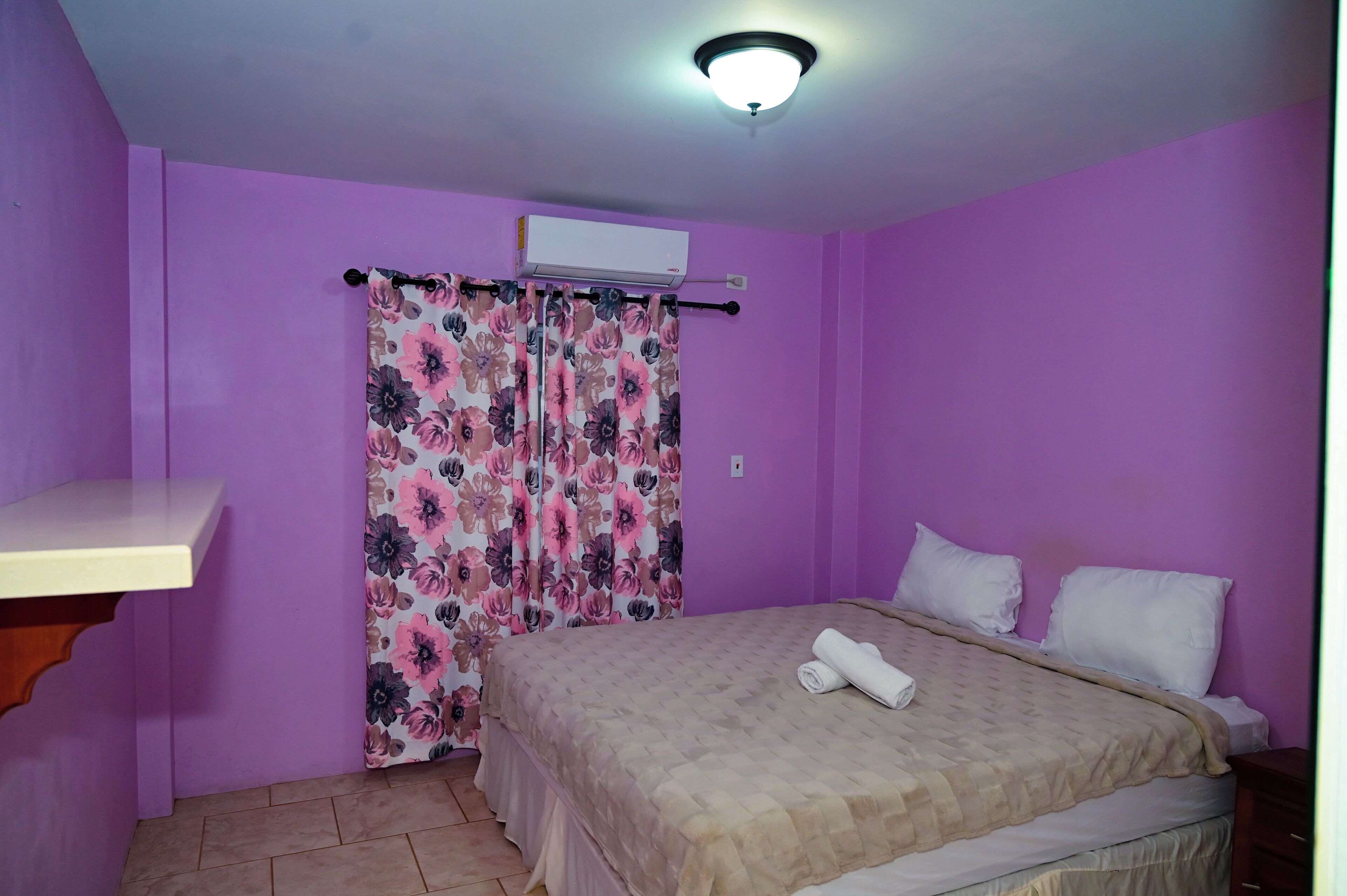 A bedroom with a bed, a nightstand, a wall-mounted air conditioner, and a window with floral curtains.