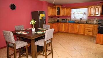 A kitchen with wooden cabinets, a dining table with chairs, a refrigerator, a microwave, and a clock on the wall.
