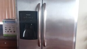 A stainless steel refrigerator with a water dispenser, located in a kitchen with wooden cabinets and a wooden floor.