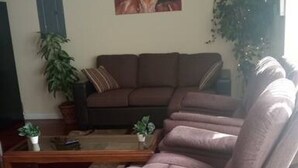 A living room with a brown sofa, a brown recliner, and a glass coffee table. There are two potted plants and a painting on the wall.
