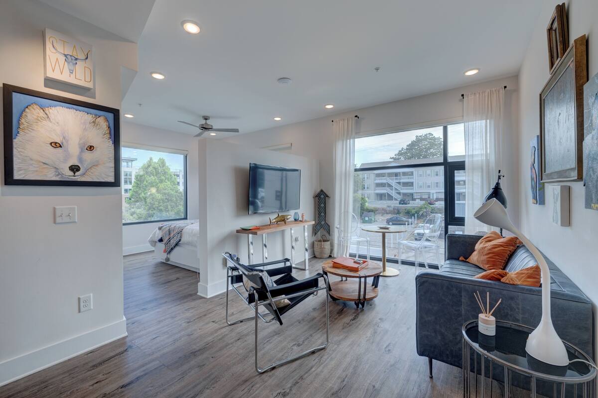 Un salon moderne avec un canapé, une table basse et un fauteuil. Un téléviseur est fixé au mur, il y a un ventilateur de plafond et une fenêtre donnant sur les bâtiments à l’extérieur.
