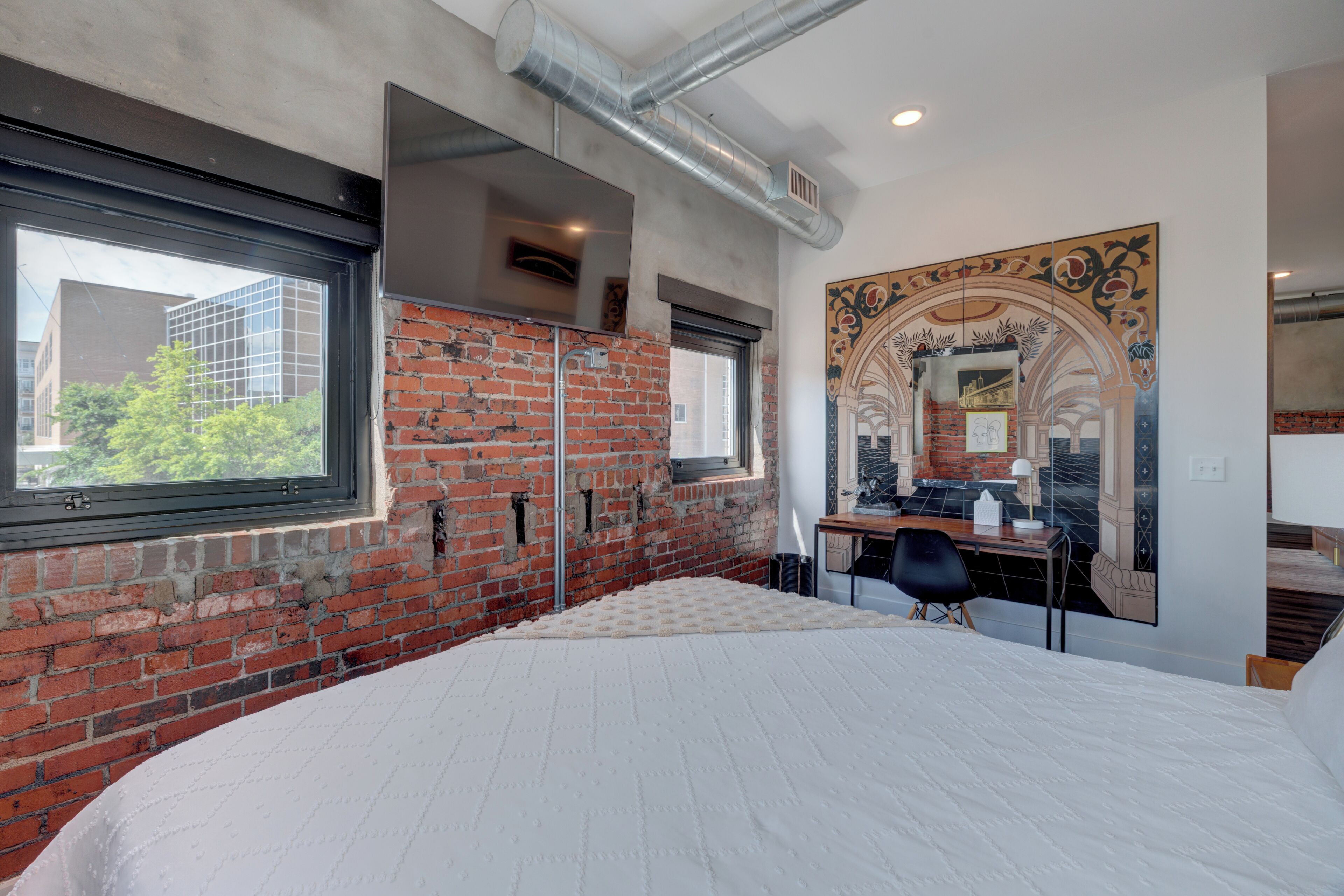 A bedroom with a brick wall, a large window, a bed, a desk, and a decorative mirror.
