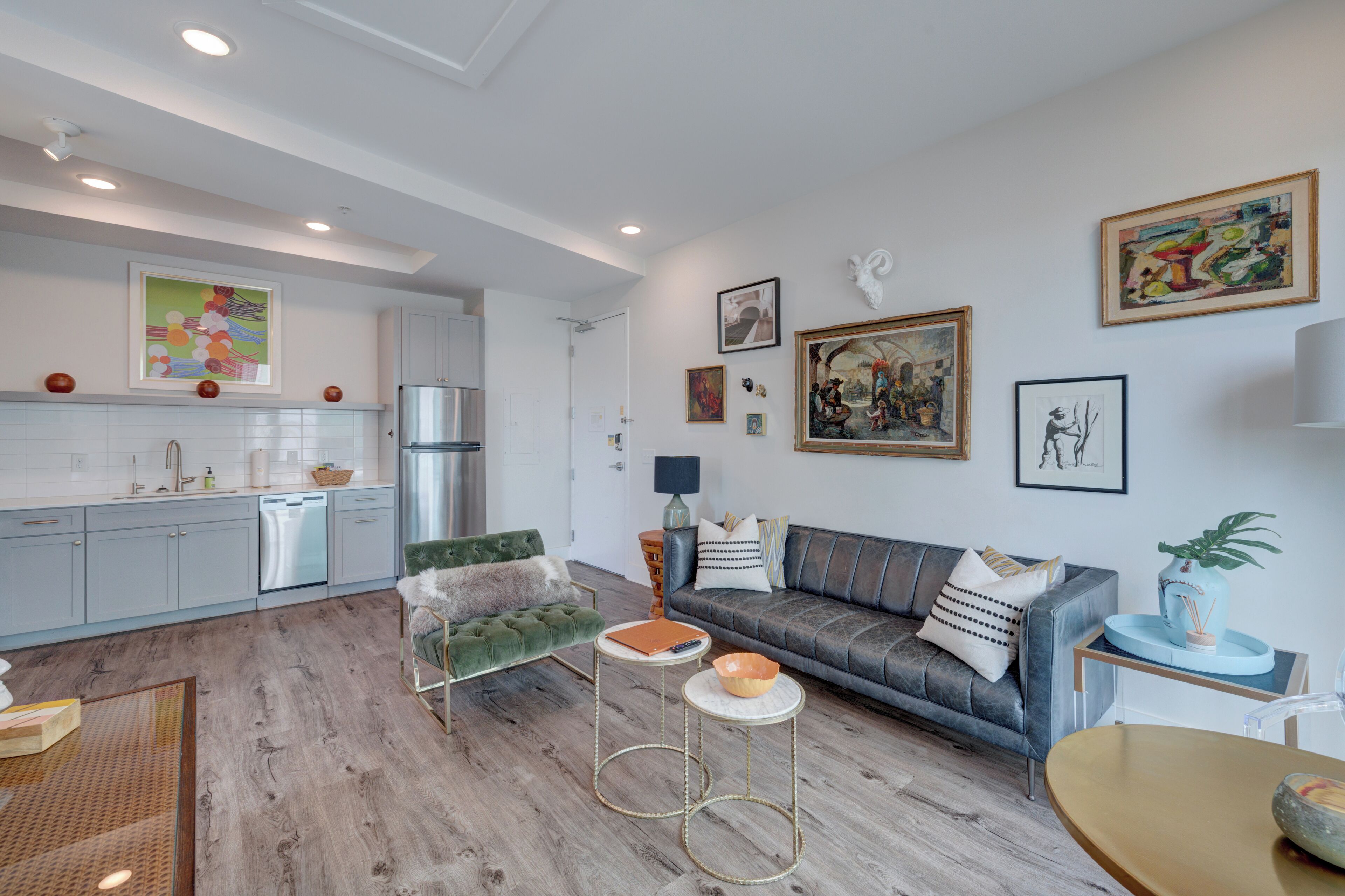 Una sala de estar moderna con un sofá gris, un sillón verde y una mesa de centro. Hay una zona de cocina con armarios blancos y un lavavajillas. El ambiente tiene suelo de madera y presenta obras de arte en las paredes.