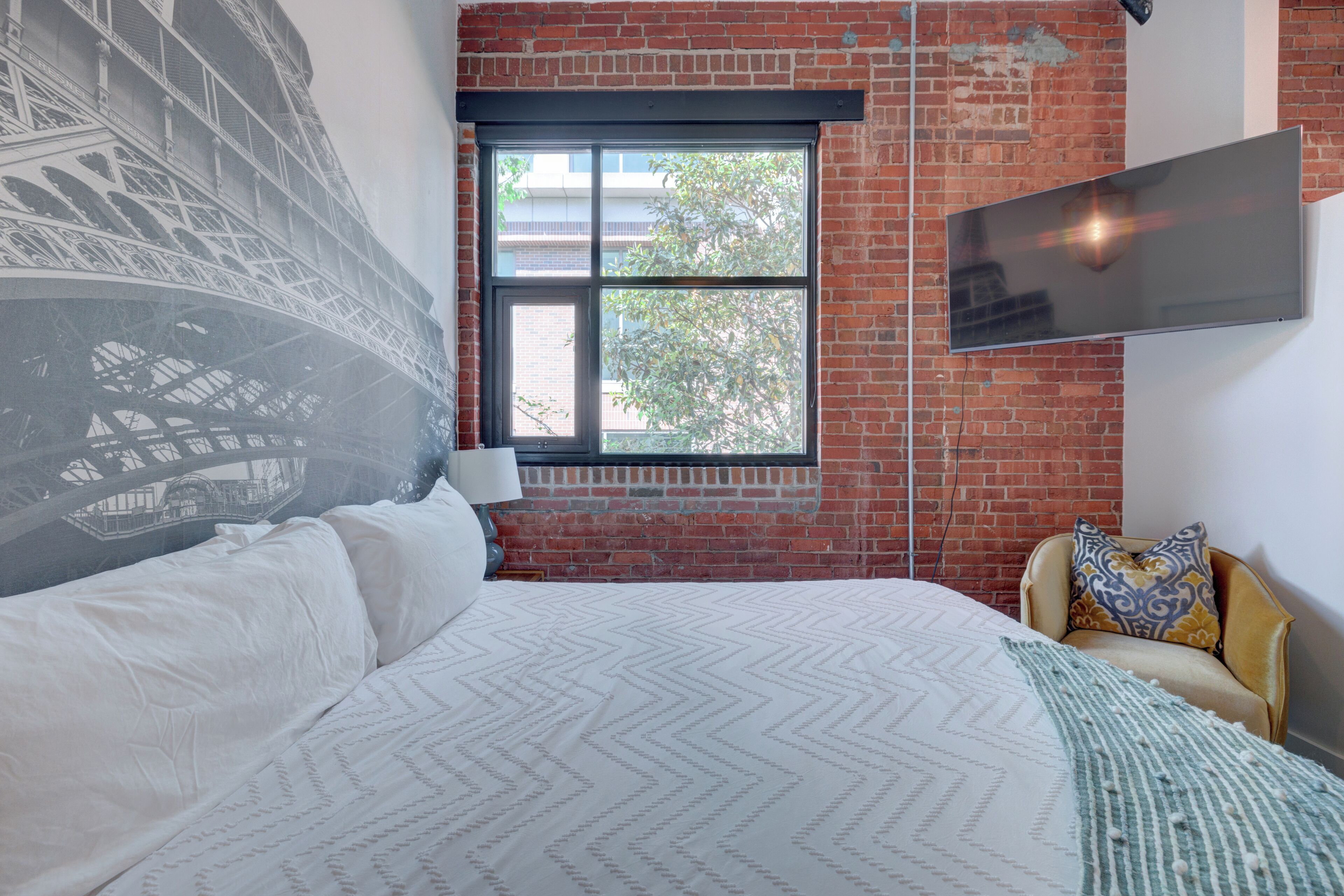 A bedroom with a large bed, a window with a view of a building, a chair with a patterned cushion, and a flat-screen TV mounted on the wall.