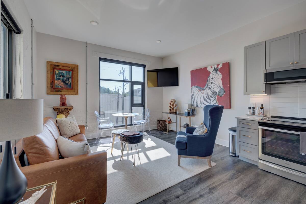 Un salon moderne avec un canapé, une table basse et des fauteuils, une cuisine équipée d’une cuisinière et de placards, et une grande fenêtre.