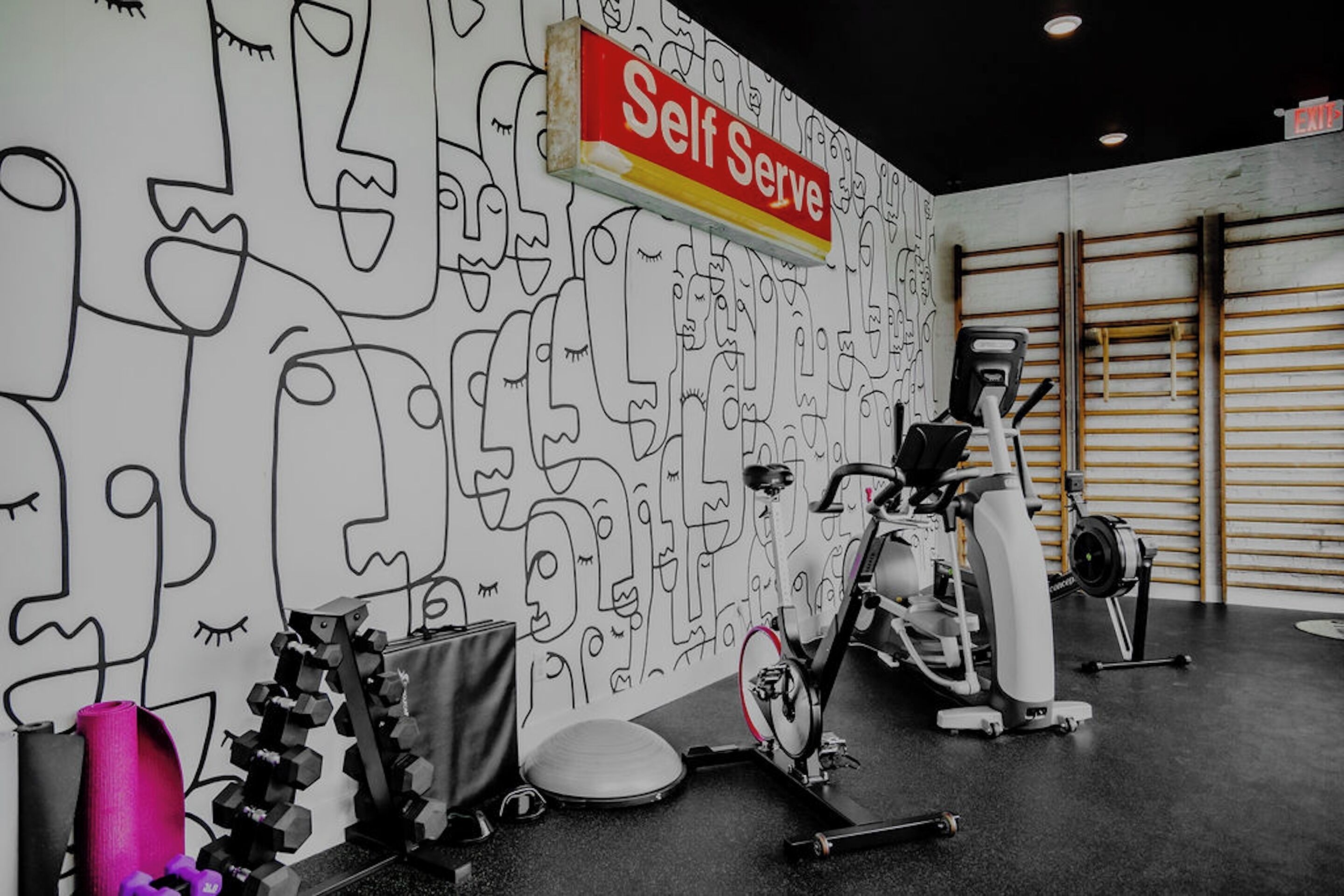 A gym with a wall of abstract line drawings, a stationary bike, dumbbells, and a yoga mat.