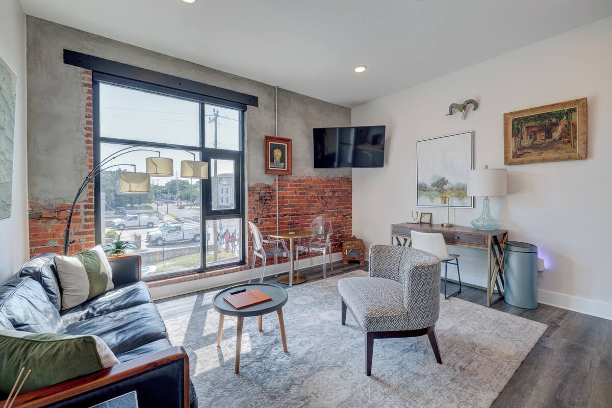 Un salon moderne avec une grande fenêtre, un canapé, un fauteuil à motifs, une petite table et un bureau avec une lampe.