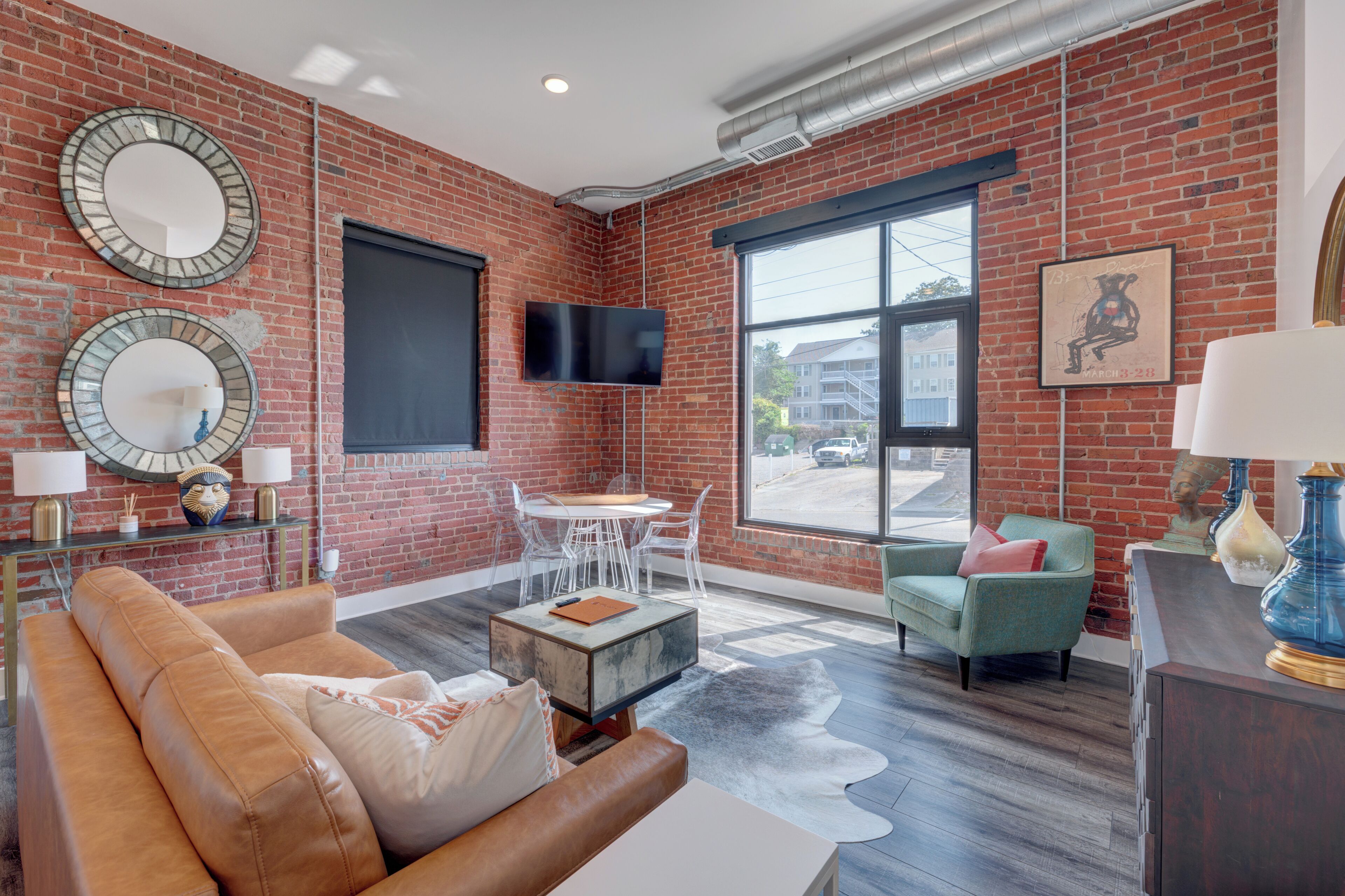 A modern living room with a brick wall, a flat-screen TV, a dining area with a table and chairs, and a large window with a view.