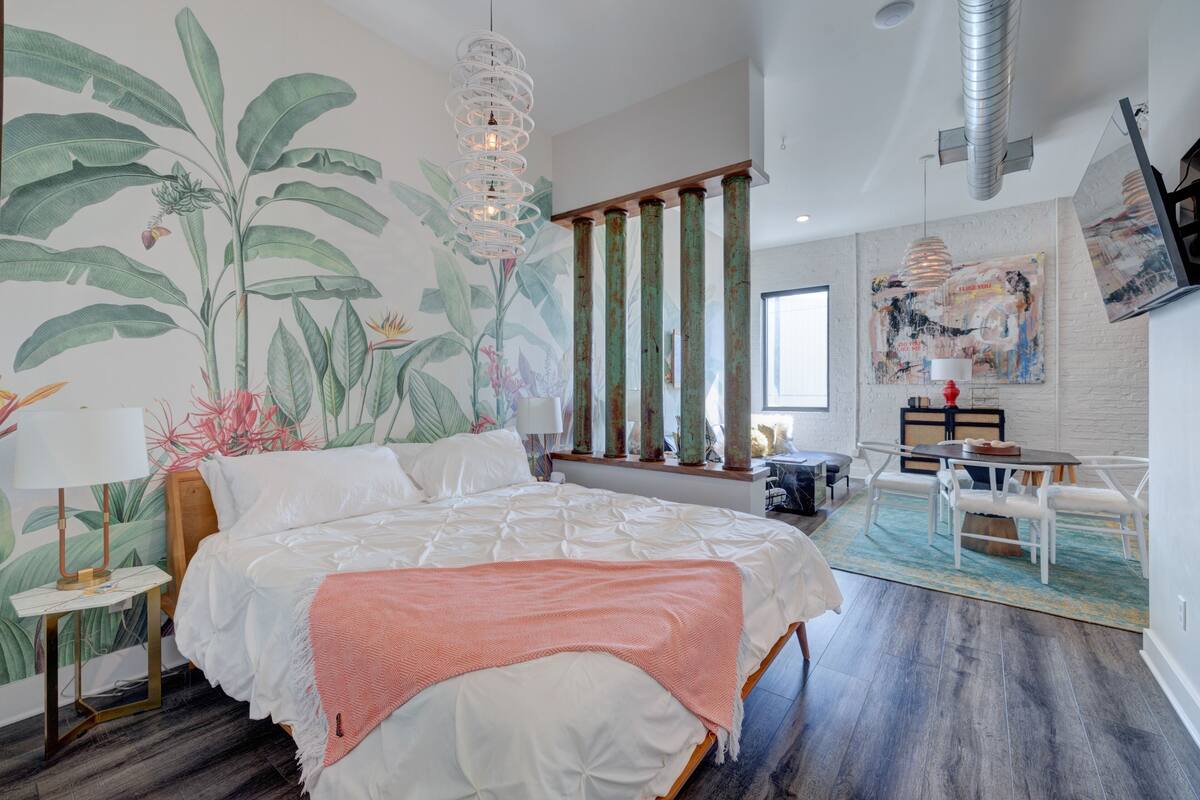 Une chambre avec un grand lit, une table de chevet, un bureau et une chaise. Il y a une grande fenêtre et un tableau au mur.
