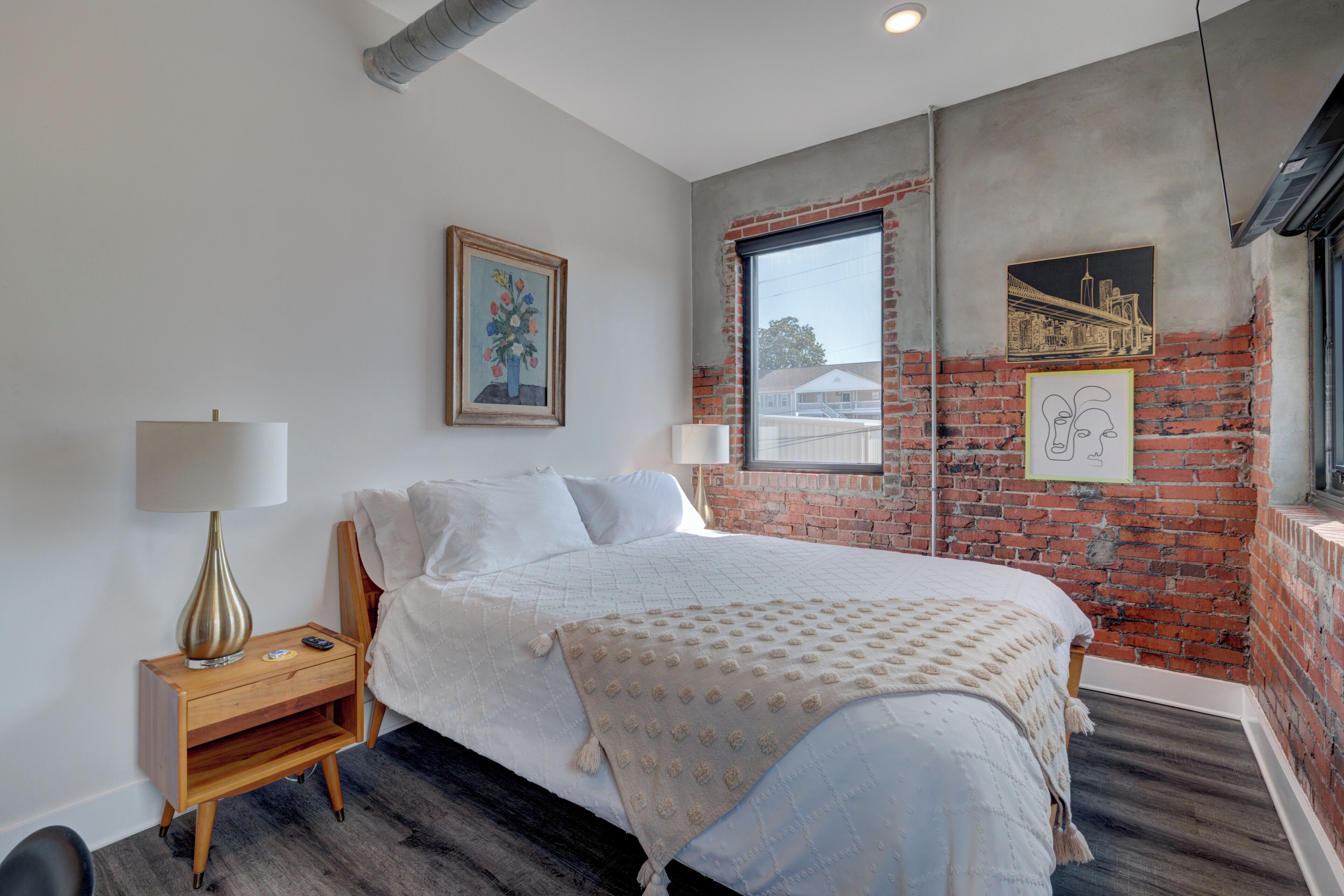 A bedroom with a bed, a nightstand, a lamp, and a window with a view of buildings.