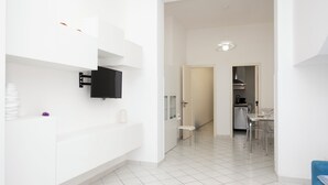 A modern, minimalist interior with a white color scheme, a flat-screen TV mounted on the wall, a small seating area, and a kitchen visible through an open door. - Eva Apartment, a stone's throw from the sea (San Vito Lo Capo)