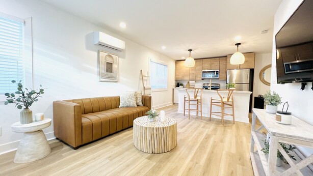 Una sala de estar moderna con un sofá de cuero marrón, una mesa de centro redonda y una cocina con armarios de madera y electrodomésticos de acero inoxidable.