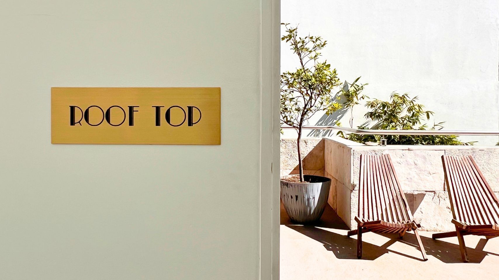 A sign labeled "ROOF TOP" and two wooden lounge chairs on an outdoor terrace with a potted plant and a wall in the background.