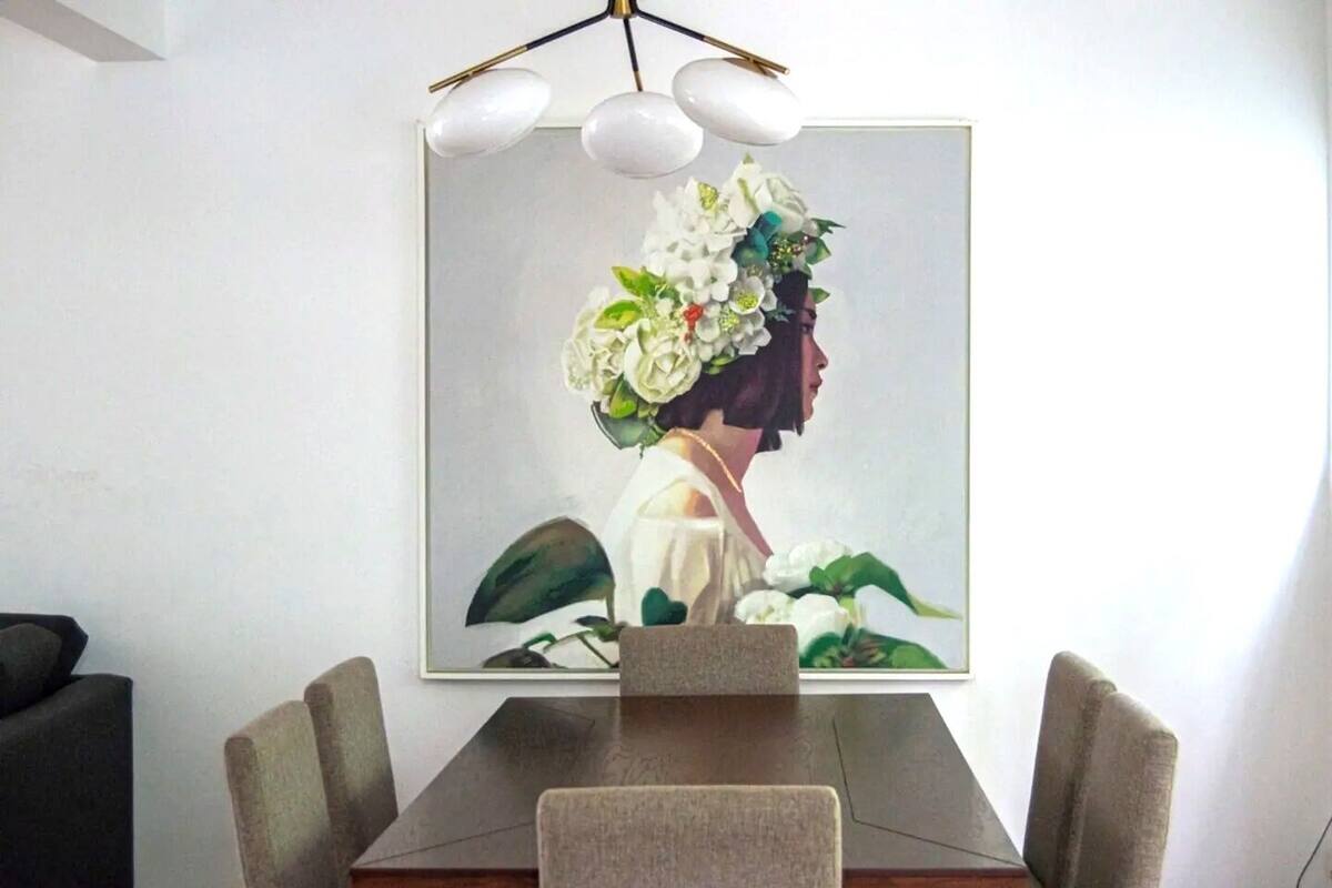 Een eetgedeelte met een rechthoekige tafel, vier stoelen en een groot schilderij van een vrouw met bloemen aan de muur.