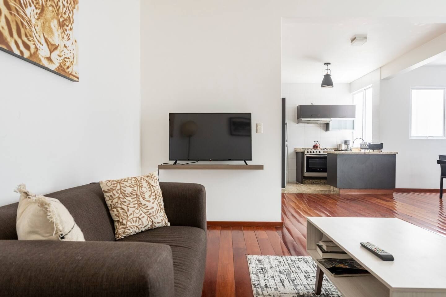 Una sala de estar moderna con sofá, televisor de pantalla plana en la pared, zona de cocina con estufa y horno, y una mesa de comedor con un control remoto.