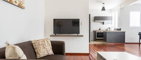 Una sala de estar moderna con sofá, televisor de pantalla plana en la pared, zona de cocina con estufa y horno, y una mesa de comedor con un control remoto.