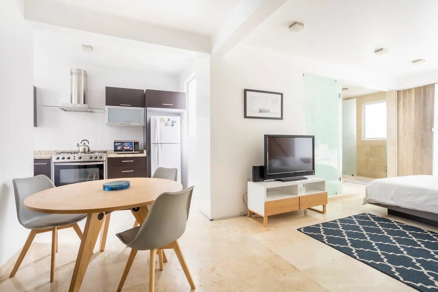 A modern hotel room with a dining area, kitchenette, TV, and a bed.