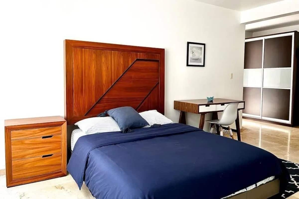 Een slaapkamer met een houten hoofdbord, een bed met blauw beddengoed, een klein houten dressoir, een bureau met stoel, en een inloopkast met schuifdeuren.