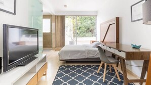 Habitación de hotel moderna con cama, escritorio, televisión y un amplio ventanal con vistas a zonas verdes.