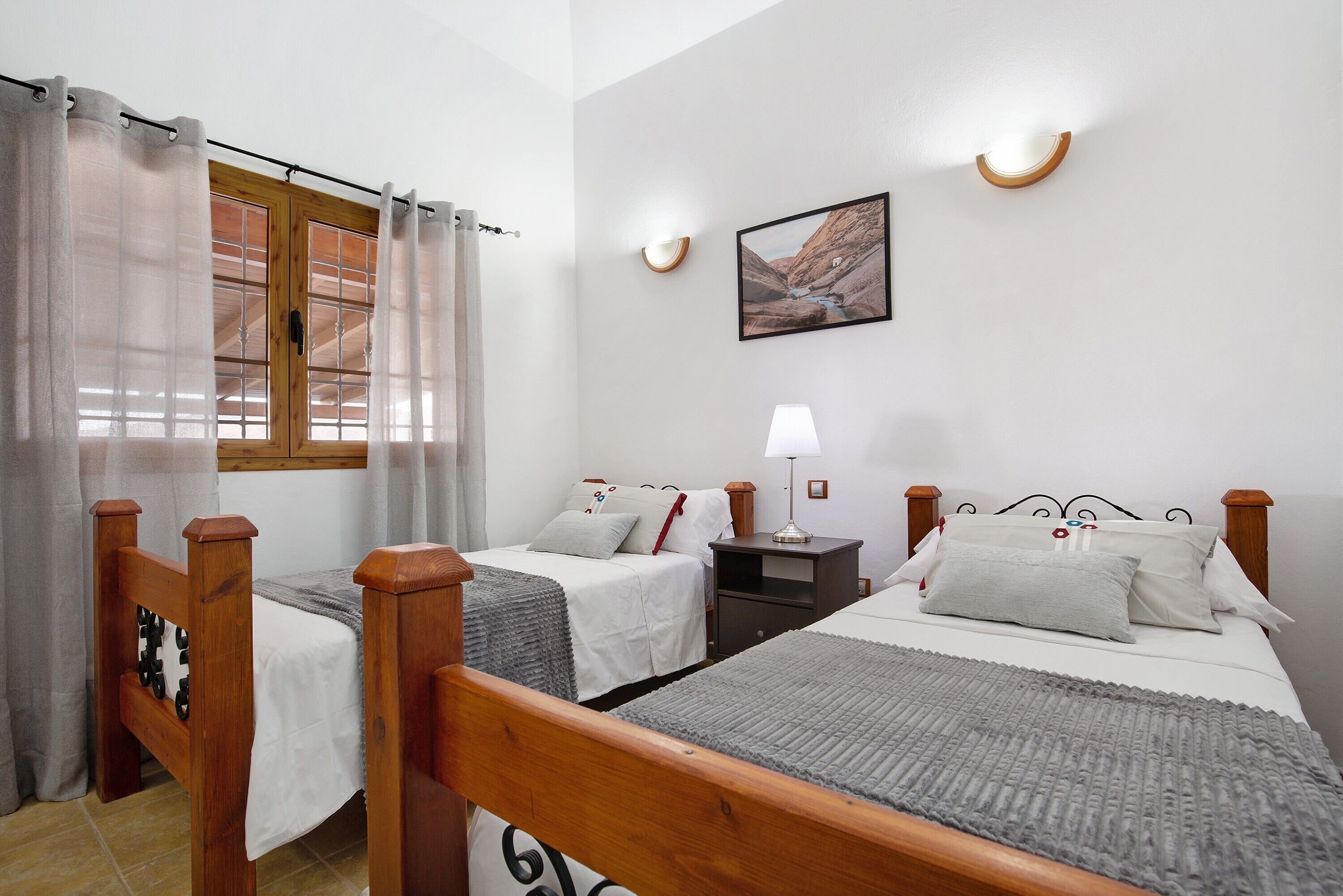 Een slaapkamer met twee bedden, een raam met gordijnen, een nachtkastje met een lamp en een aan de muur hangend schilderij.