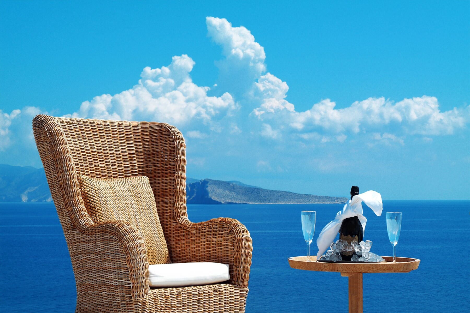 Una silla de mimbre con cojín blanco, una mesita de madera con una botella y dos vasos, y una vista panorámica del mar y las montañas.