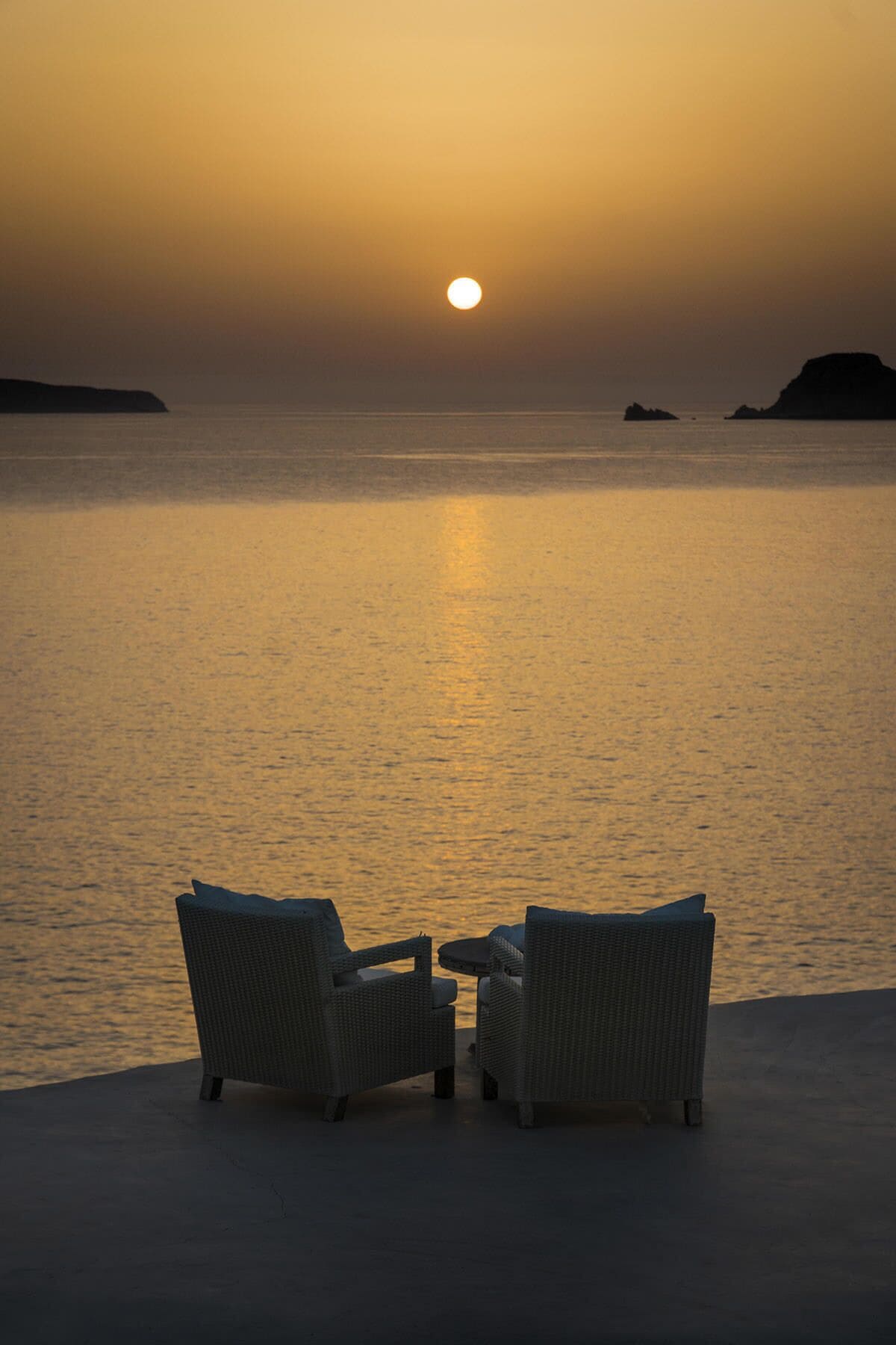 Two wicker chairs facing a calm sea at sunset.