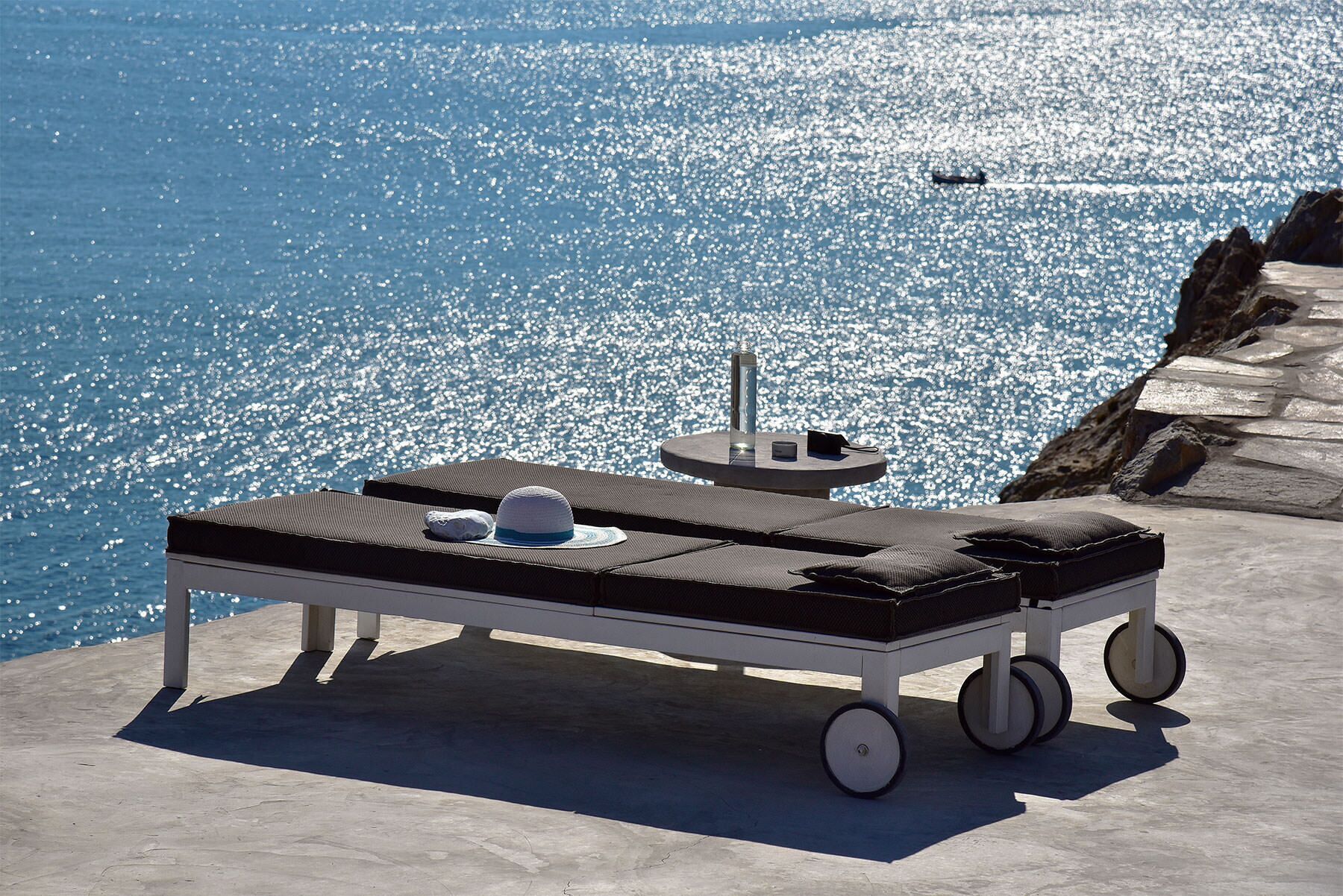 A sun lounger with a hat and a bottle on a concrete surface near a body of water.