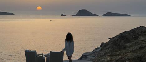 Eine Person geht an einer felsigen Küstenlinie entlang, mit Blick auf das Meer und entfernte Inseln bei Sonnenuntergang.