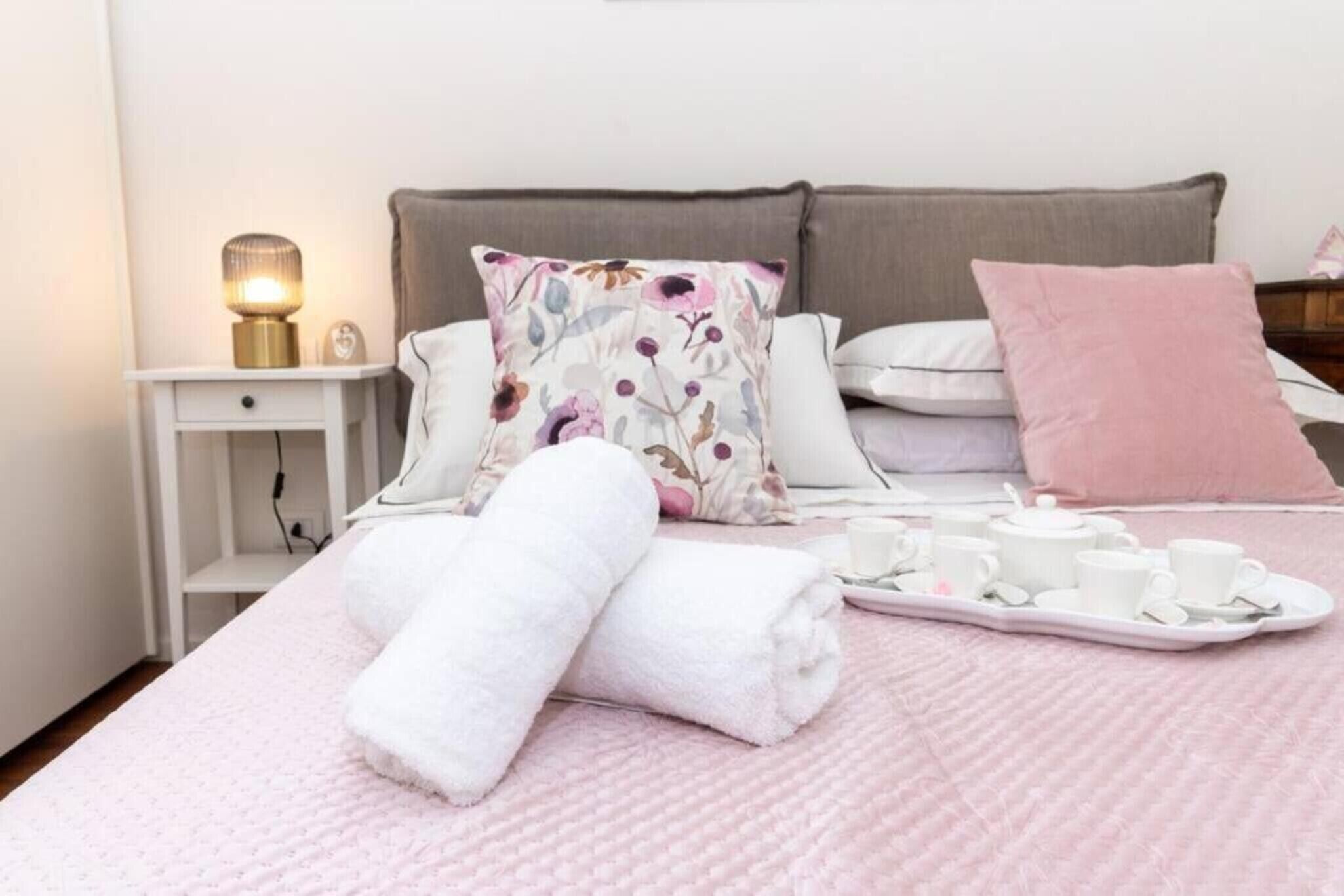 Een net opgemaakt bed met een roze quilt, witte kussens en een roze plaid. Op het bed staat een dienblad met theekopjes en een theepot.