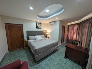 Habitación de hotel con cama, escritorio, silla, puerta, equipo de aire acondicionado y un cuadro en la pared.